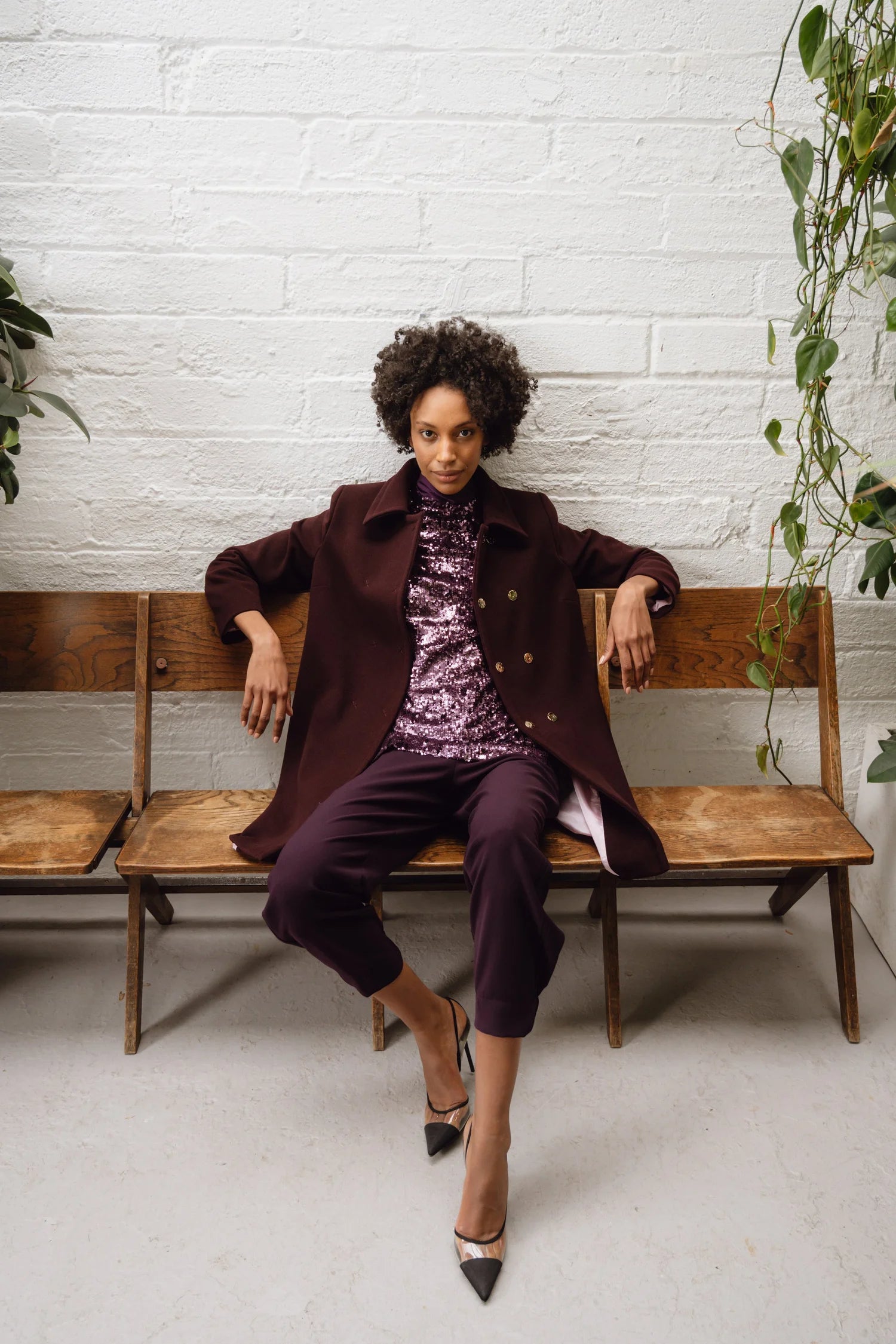 Front-facing image of woman in Havana mini skirt and wine coat, seated on wooden bench with plants in background