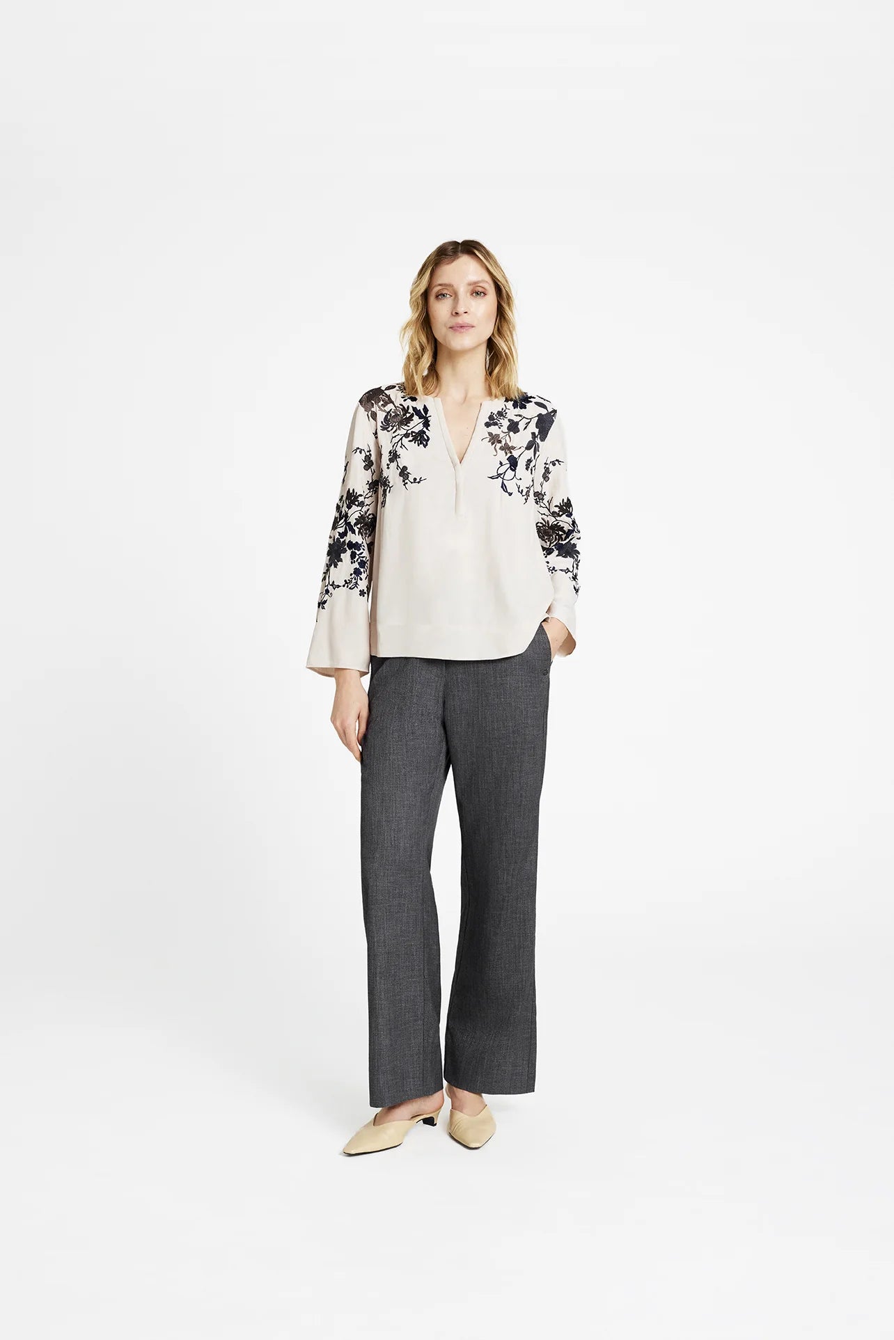 Model wearing Gustav Naja ivory blouse with navy floral embroidery, styled with grey trousers.
