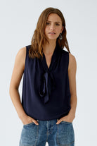 Model in navy sleeveless top, hands in pockets, soft bow at neckline