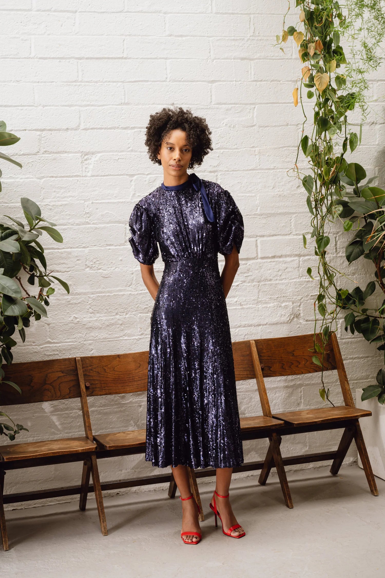 Model standing against a white brick wall in sparkly navy balloon sleeve midi dress from Sisters by Caroline Kilkenny 